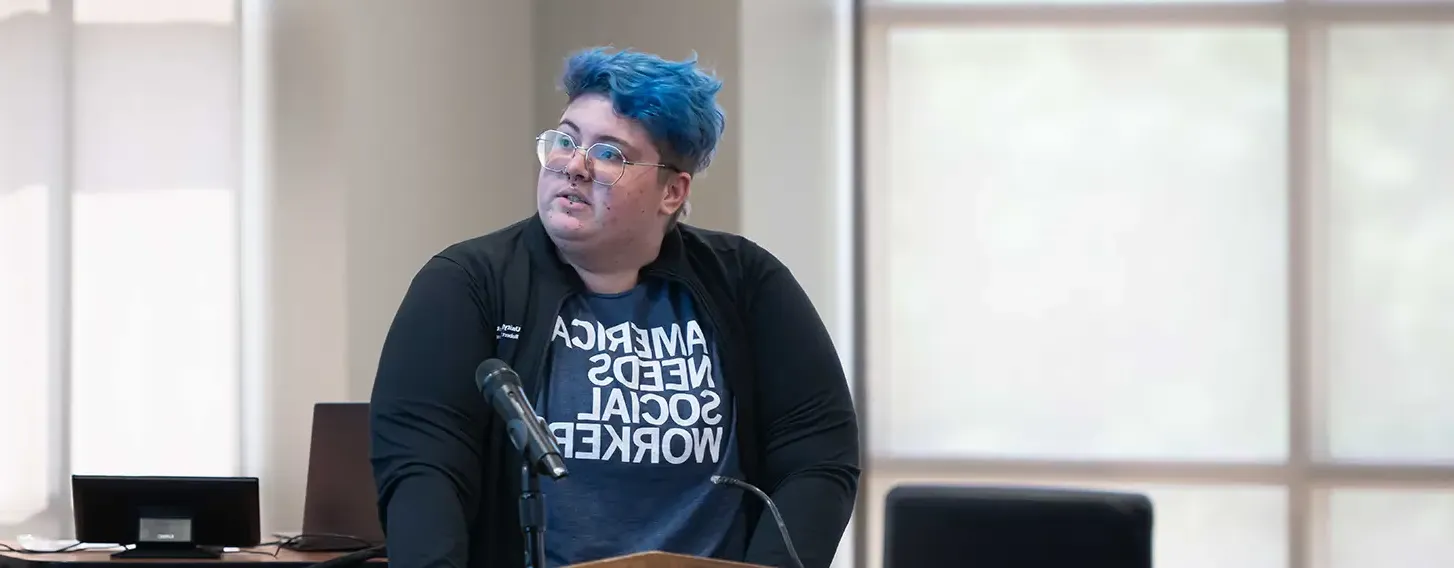 A person with blue hair standing at a podium, addressing an audience.